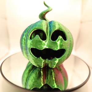 Op de afbeelding: Een groene en rode decoratieve pompoen met een uitgesneden gezicht, op een zwarte, ronde schaal. De pompoen heeft een krullende groene stengel en een glanzende afwerking, ideaal voor Halloween.