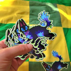 May include: A hand holds a stack of stickers featuring a stylized black cat with blue and purple accents. The cat has large blue eyes and a green outline. The background is a patchwork of green and yellow squares.