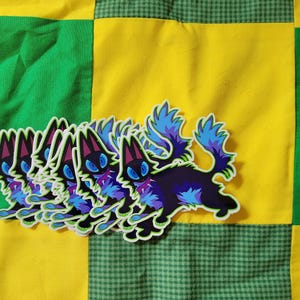 May include: A collection of stickers featuring a stylized black cat with blue and purple accents. The cat has large blue eyes and a fluffy tail. The stickers are on a yellow and green patchwork background.