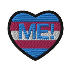 Pode incluir: Um remendo em forma de coração com um design de bandeira do orgulho transgénero. O remendo é azul, rosa e branco com a palavra "ME!" em letras azuis.