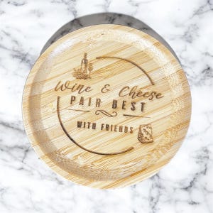 May include: A round bamboo serving tray with engraved text and illustrations. The text reads "Wine & Cheese Pair Best With Friends." Illustrations include a wine bottle, grapes, a wine glass, and a cheese wedge. The tray has a natural wood grain.