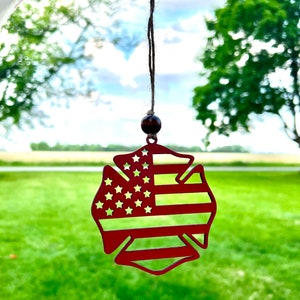 May include: Red metal firefighter's helmet ornament with a cutout of the American flag. The ornament is hanging from a brown string.