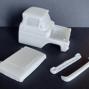 May include: A white, unassembled model truck cab with separate hood, bumper, and other parts. The model appears to be made of plastic and is designed for hobbyists or model builders. The cab features windows and a detailed front end.
