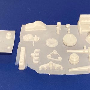 May include: A collection of white plastic model car engine parts, including an engine block, cylinders, and various other components. The parts are arranged on a blue surface, suggesting a model-making project.