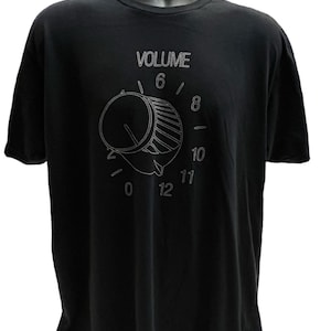 May include: Black t-shirt featuring a white graphic of a volume knob with the word "VOLUME" above it. The knob is set to the number 6. Numbers 0, 8, 10, 11, and 12 are also visible on the dial. The shirt is displayed on a black mannequin.