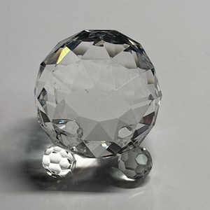 May include: A clear, faceted crystal sphere with three smaller crystal spheres as feet. The large sphere has a geometric design, reflecting light. The decorative object is set against a white background.