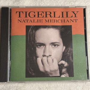 May include: CD case for the album "Tigerlily" by Natalie Merchant. The album cover features a black and white portrait of a woman with long hair, set against a background of orange and green.