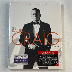 May include: The Daniel Craig Collection Blu-ray disc. The cover features a black and white image of Daniel Craig in a tuxedo holding a gun. The title is in orange and white lettering. Includes Digital HD and 7 classic James Bond theme songs.