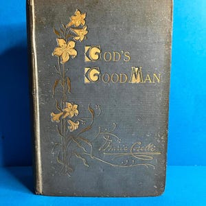 May include: Antique book titled "God's Good Man" by Marie Corelli. The cover is a faded blue-gray with gold floral accents and lettering. The book shows signs of age and wear.