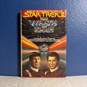 May include: A paperback book cover for the novel "Star Trek II: The Wrath of Khan" by Vonda N. McIntyre. The cover features a colorful illustration of Captain Kirk, Spock, and Khan Noonien Singh. The book is based on the Paramount Pictures film.