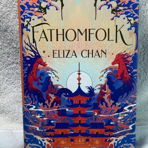 May include: A hardcover book titled "FATHOMFOLK" by Eliza Chan. The cover features a vibrant illustration with a pagoda, dragons, and a full moon. The color palette includes shades of orange, blue, and red.