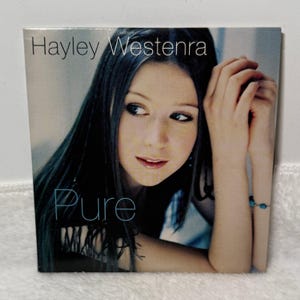 May include: A square music CD album cover featuring a portrait of Hayley Westenra with long dark hair. The text Hayley Westenra is printed in the top left corner, and the word Pure is printed in the bottom left corner in a light blue font.