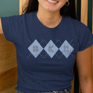 May include: A navy blue t-shirt with a light blue argyle pattern and the letters "S K C" in the center.