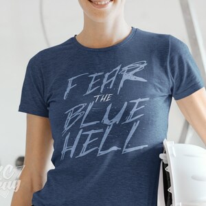 May include: A woman wearing a blue t-shirt with the text "Fear the Blue Hell" in white. She is holding a white hard hat.