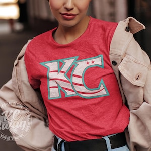 KC Swag - Kansas City Current, Flowing KC on Heather red unisex t-shirt worn by female model under an open jacket standing in front of an old barn