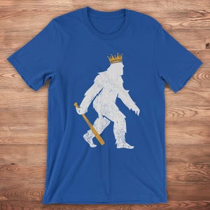 shopkcswag, a traditional royal blue kansas city royals pasquantino t-shirt, featuring a distressed white silhouette of a strutting "pasquatch" donning a gold crown and dragging a gold bat, shown on a wood surface
