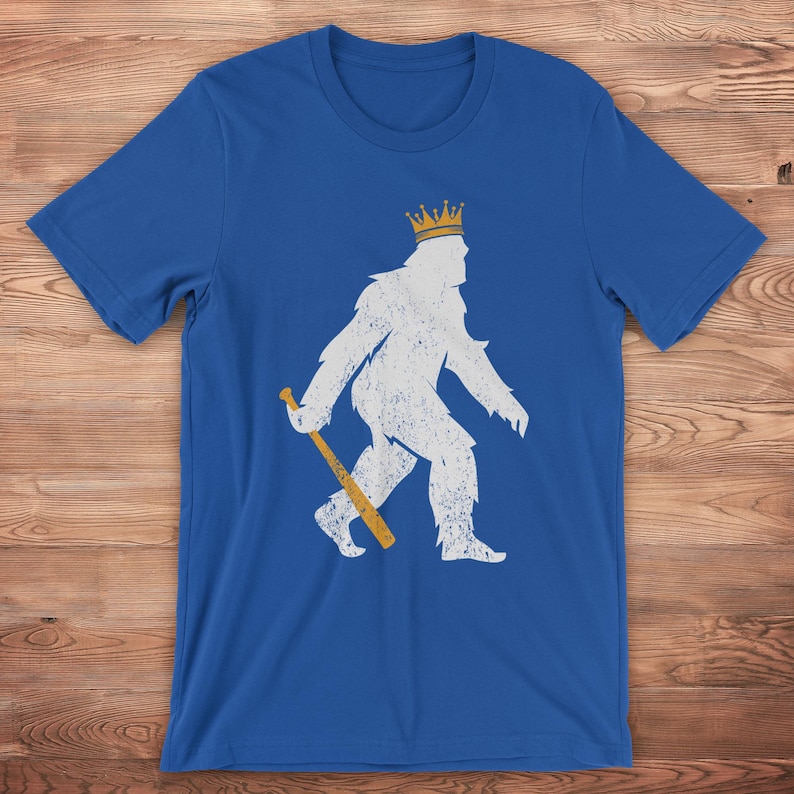 shopkcswag, a traditional royal blue kansas city royals pasquantino t-shirt, featuring a distressed white silhouette of a strutting "pasquatch" donning a gold crown and dragging a gold bat, shown on a wood surface