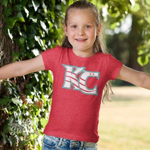 KC Swag - Kansas City Current, Flowing KC on Heather red unisex t-shirt worn by youth girl model standing between trees in a public park