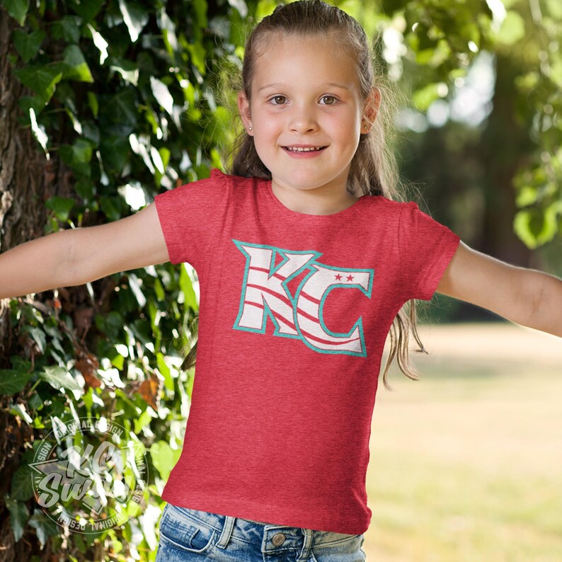 KC Swag - Kansas City Current, Flowing KC on Heather red unisex t-shirt worn by youth girl model standing between trees in a public park