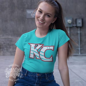 KC Swag - Kansas City Current, Flowing KC on Teal unisex t-shirt worn by female model sitting in a concrete alley
