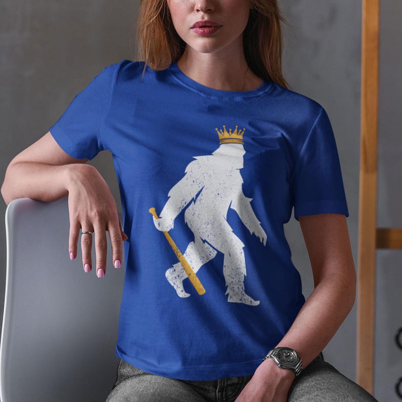 shopkcswag, female model sitting in casual indoor lifestyle setting, wearing a traditional royal blue kansas city royals pasquantino t-shirt, featuring a distressed white silhouette of a strutting "pasquatch" donning a gold crown and dragging a gold bat