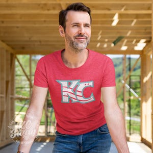 KC Swag - Kansas City Current, Flowing KC on Heather red unisex t-shirt worn by male model standing in a framed home construction zone