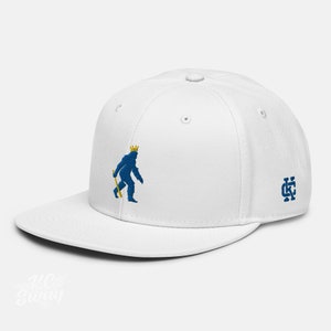 May include: White baseball cap with a blue embroidered image of a sasquatch wearing a gold crown and holding a stick. The cap has a blue embroidered "KC" logo on the right side.