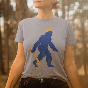 shopkcswag, female model standing in casual outdoor lifestyle setting, wearing a classic heather gray kansas city royals pasquantino t-shirt, featuring a distressed royal blue silhouette of a strutting "pasquatch" donning a gold crown and dragging a gold bat