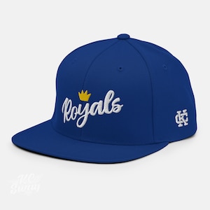 Puede incluir: Una gorra de béisbol azul real con una corona bordada en blanco y la palabra "Royals" en letras blancas cursivas. La gorra también tiene un pequeño "KC" bordado en blanco en el lado derecho.