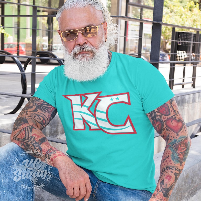 KC Swag - Kansas City Current, Flowing KC on Teal unisex t-shirt worn by a bearded and tattooed male model sitting in an outdoor plaza