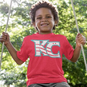 KC Swag - Kansas City Current, Flowing KC on Heather red unisex t-shirt worn by youth boy model sitting on a swing in a public park