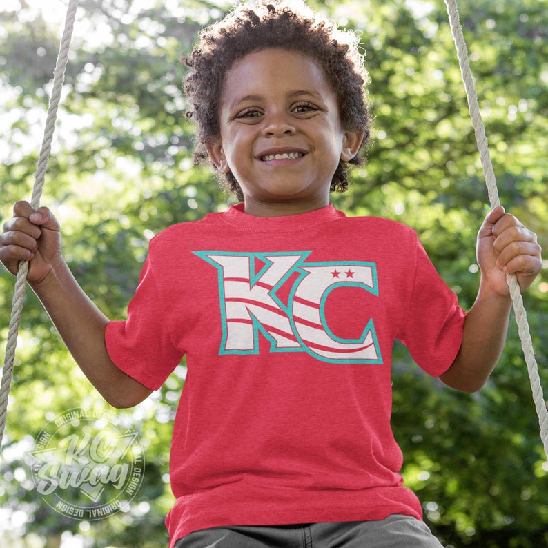 KC Swag - Kansas City Current, Flowing KC on Heather red unisex t-shirt worn by youth boy model sitting on a swing in a public park