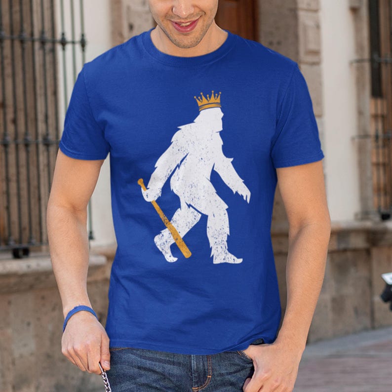 shopkcswag, male model standing in casual outdoor lifestyle setting, wearing a traditional royal blue kansas city royals pasquantino t-shirt, featuring a distressed white silhouette of a strutting "pasquatch" donning a gold crown and dragging a gold bat