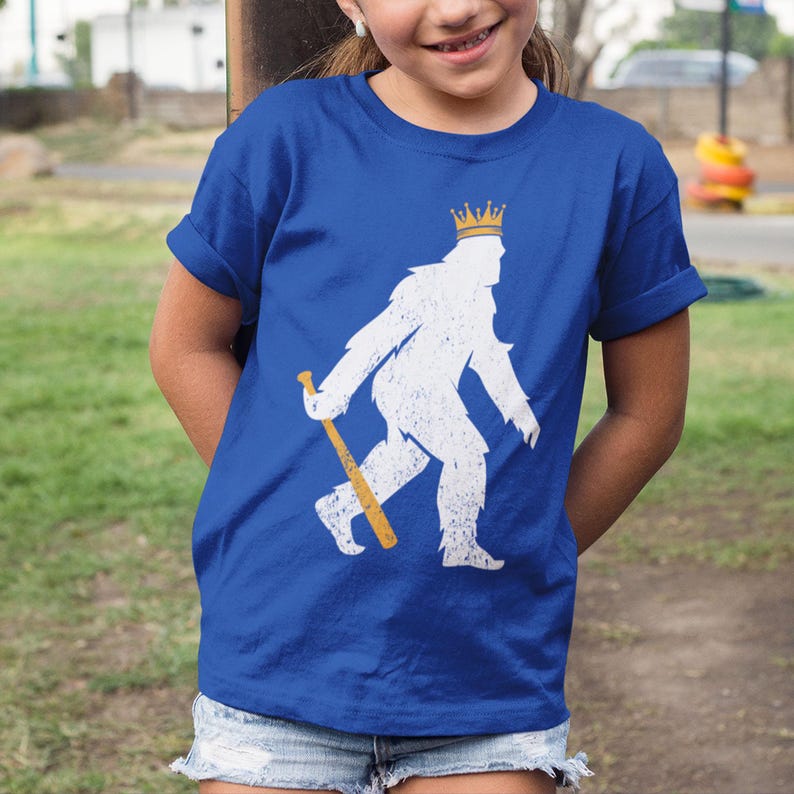 shopkcswag, youth girl model sitting in casual outdoor lifestyle setting, wearing a traditional royal blue kansas city royals pasquantino t-shirt, featuring a distressed white silhouette of a strutting "pasquatch" donning a gold crown and dragging a gold bat