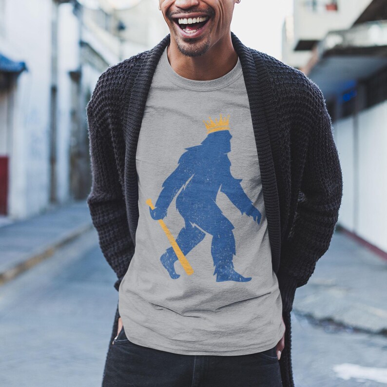 shopkcswag, male model standing in casual outdoor urban lifestyle setting, wearing a classic heather gray kansas city royals pasquantino t-shirt, featuring a distressed royal blue silhouette of a strutting "pasquatch" donning a gold crown and dragging a gold bat