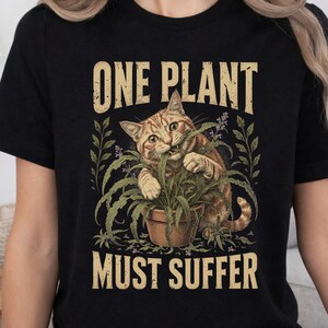 Puede incluir: Una camiseta negra con el gráfico de un gato atigrado naranja mordiendo una planta verde en una maceta marrón. El texto sobre el gato dice ONE PLANT y el texto debajo dice MUST SUFFER en una tipografía clara con un efecto desgastado.