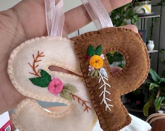 May include: Handmade felt letter ornaments, featuring the letters "S" and "P". The "S" is cream-colored, and the "P" is brown. Both letters are adorned with embroidered floral and leaf details, and have pink ribbon hangers.