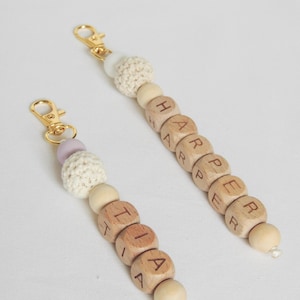 Può includere: Due portachiavi con perline di legno con i nomi "Harper" e "Tia" scritti sulle perline. Le perline sono infilate su un cordoncino con un moschettone in metallo dorato. Le perline sono di colore marrone chiaro e hanno una perlina di uncinetto bianca in cima.