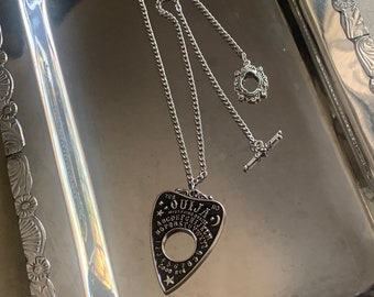 Könnte beinhalten: Eine silberne Halskette mit einem silbernen Ouija-Brett-Anhänger. Der Anhänger ist tropfenförmig und hat die Buchstaben des Alphabets, Zahlen und die Wörter "ja", "nein" und "auf Wiedersehen" darauf.