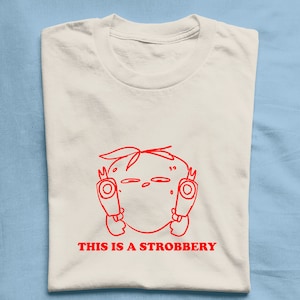 May include: White t-shirt with a red line drawing of a strawberry with arms and legs holding a gun in each hand. The text "THIS IS A STROBBBERY" is printed below the image.