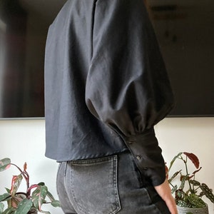 May include: A black blouse with long sleeves and a slightly cropped hem. The sleeves have a gathered, puffed look at the shoulders and are fitted at the wrists with button closures.