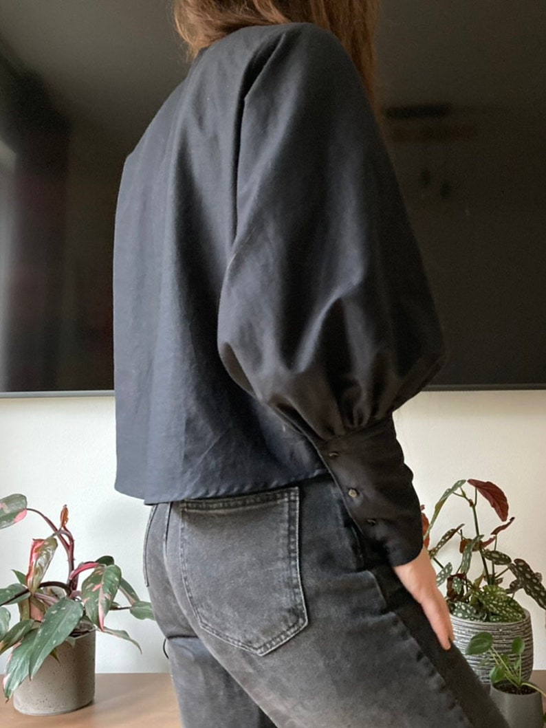 May include: A black blouse with long sleeves and a slightly cropped hem. The sleeves have a gathered, puffed look at the shoulders and are fitted at the wrists with button closures.