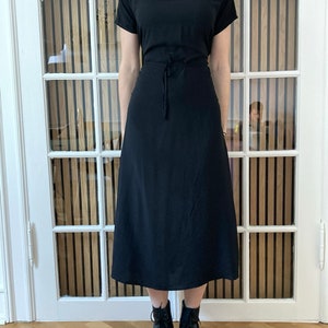 May include: A black midi dress with a mock neck and short sleeves. The dress has a tie at the waist and falls to the mid-calf. The model is wearing black ankle boots.