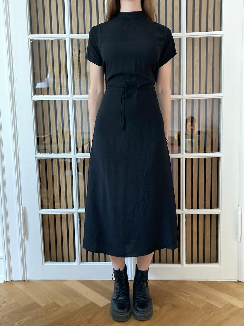 May include: A black midi dress with a mock neck and short sleeves. The dress has a tie at the waist and falls to the mid-calf. The model is wearing black ankle boots.