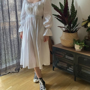 May include: A white, long-sleeved dress with a square neckline and a gathered waist. The dress has a flowing skirt and is made of a lightweight fabric. The model is wearing black high-top sneakers.