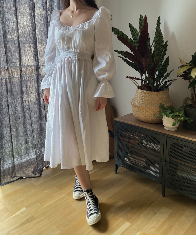 May include: A white, long-sleeved dress with a square neckline and a gathered waist. The dress has a flowing skirt and is made of a lightweight fabric. The model is wearing black high-top sneakers.