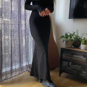 K&ouml;nnte beinhalten: Ein schwarzes, schulterfreies Maxikleid mit langen &Auml;rmeln. Das Kleid hat einen figurbetonten Oberteil und einen flie&szlig;enden Rock. Das Model tr&auml;gt schwarze Plateauschuhe.