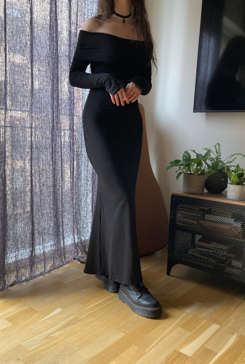 K&ouml;nnte beinhalten: Ein schwarzes, schulterfreies Maxikleid mit langen &Auml;rmeln. Das Kleid hat einen figurbetonten Oberteil und einen flie&szlig;enden Rock. Das Model tr&auml;gt schwarze Plateauschuhe.