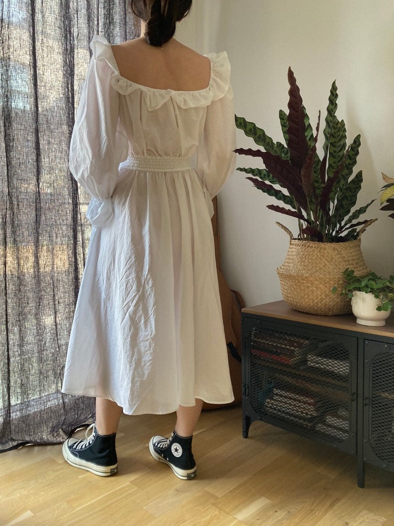 May include: A white, long-sleeved dress with a ruffled neckline and a cinched waist. The dress is made of a lightweight fabric and has a flowy silhouette. The model is wearing black high-top sneakers.