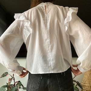 May include: White long-sleeve blouse with a ruffled neckline and puffed sleeves. The blouse has a relaxed fit and a slightly cropped hem.